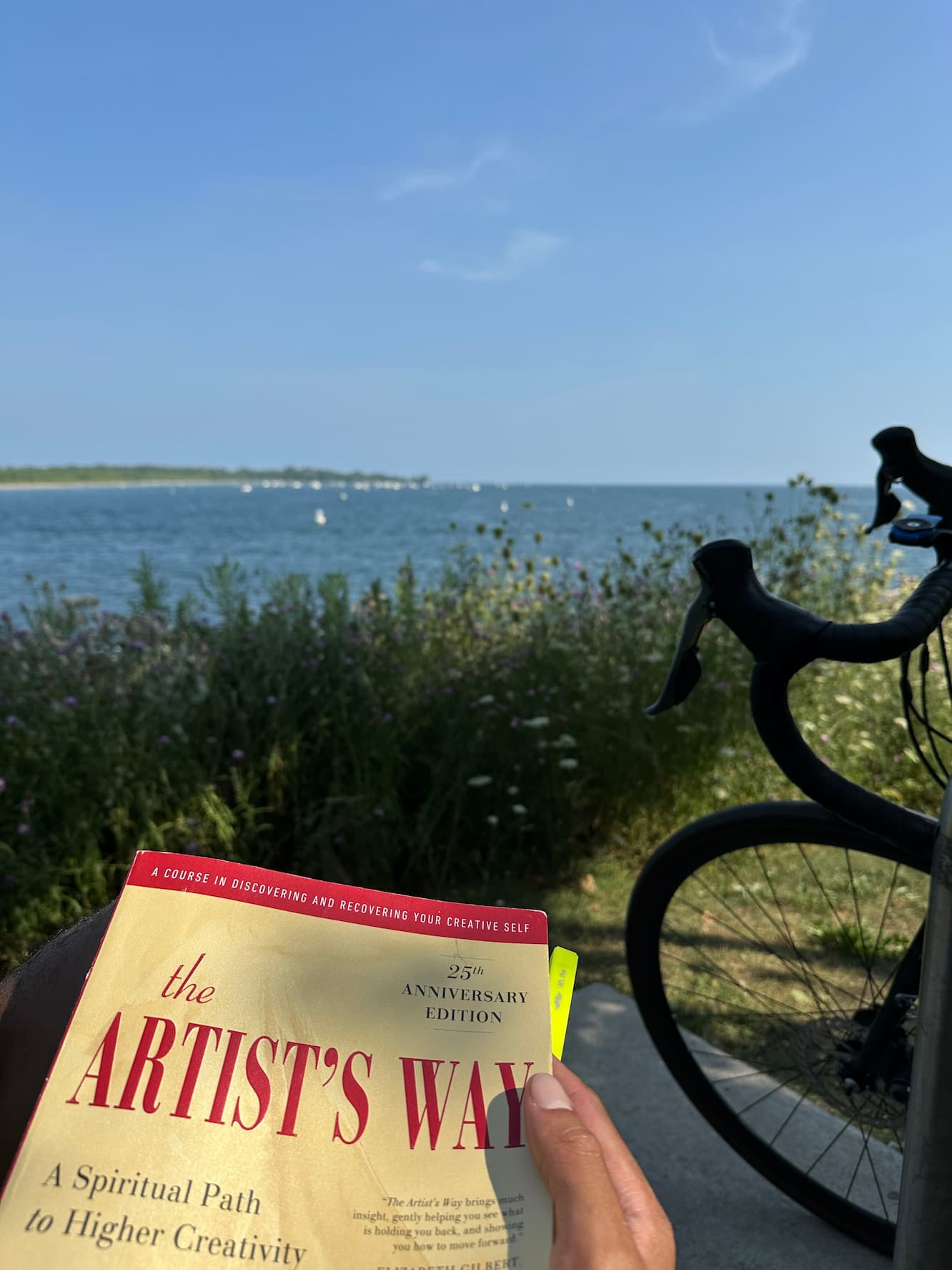 A hand holding The Artist's Way book in the foreground, with a bicycle wheel visible on the right and a scenic view of blue water, distant shoreline, and sky with scattered clouds in the background.