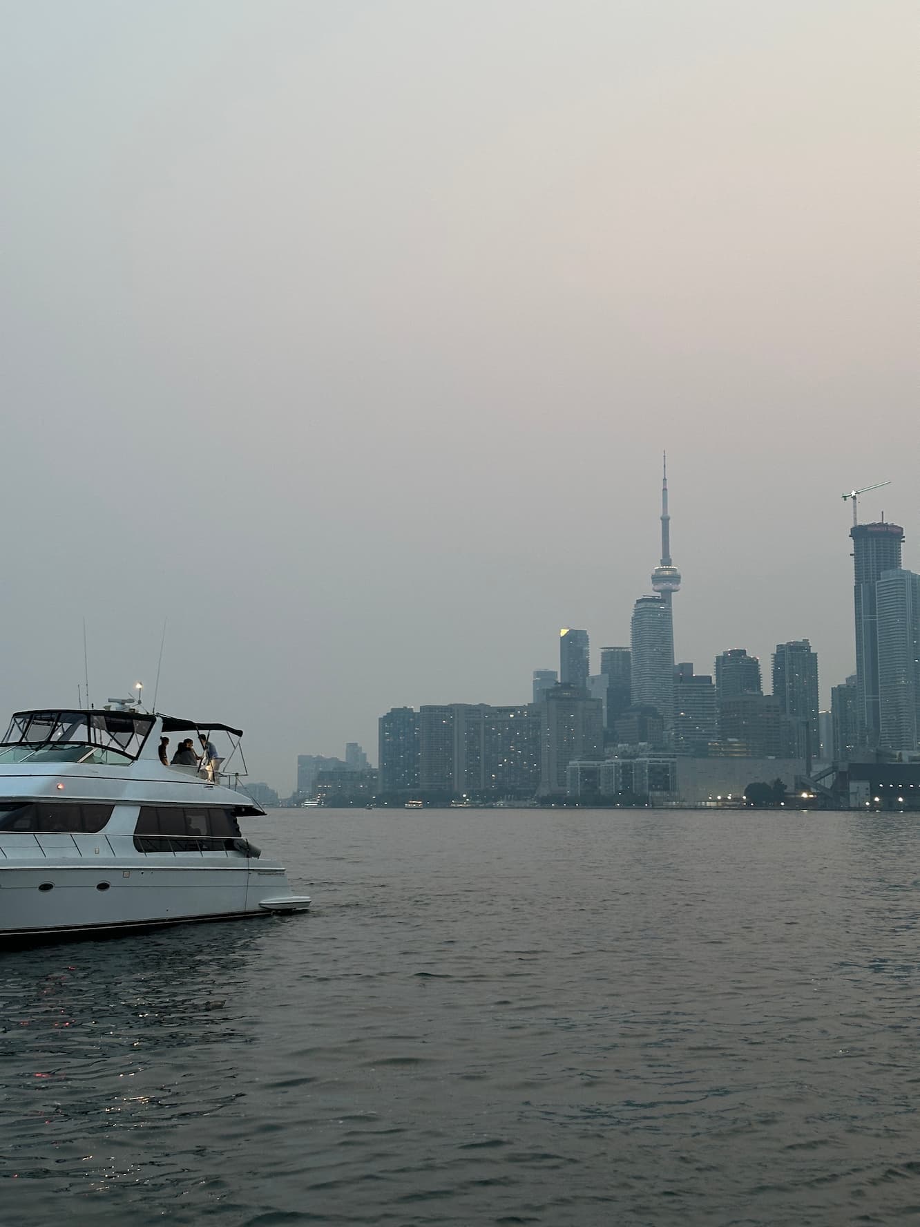 Yacht on the water at dusk with Toronto skyline and the CN Tower in a hazy sky.