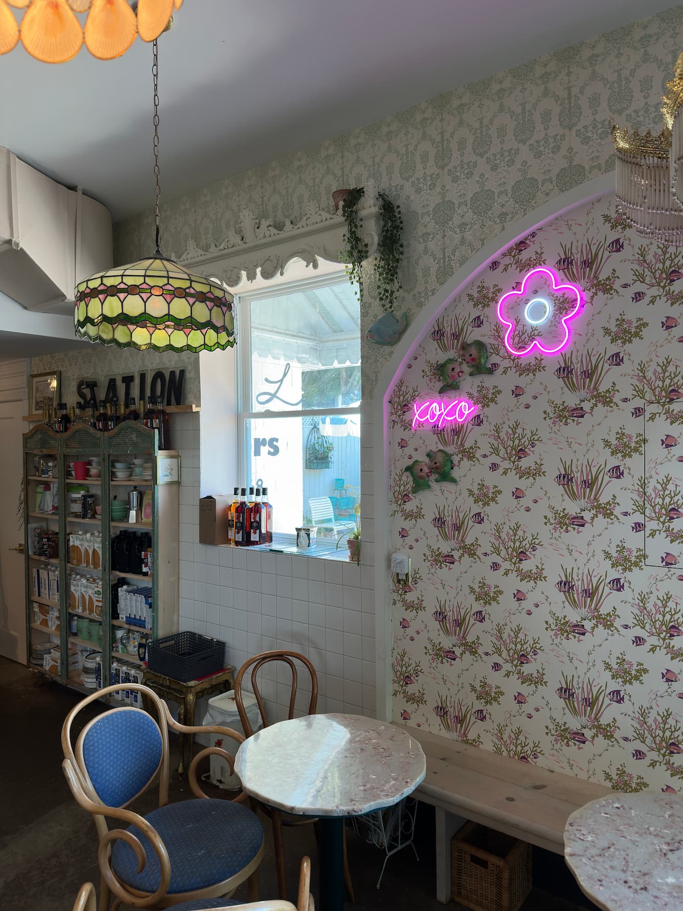 A cozy cafe interior with floral wallpaper, marble tables, bentwood chairs with blue upholstery, a vintage pendant lamp, pink neon signs (including XOXO), and shelving displaying products along the walls.