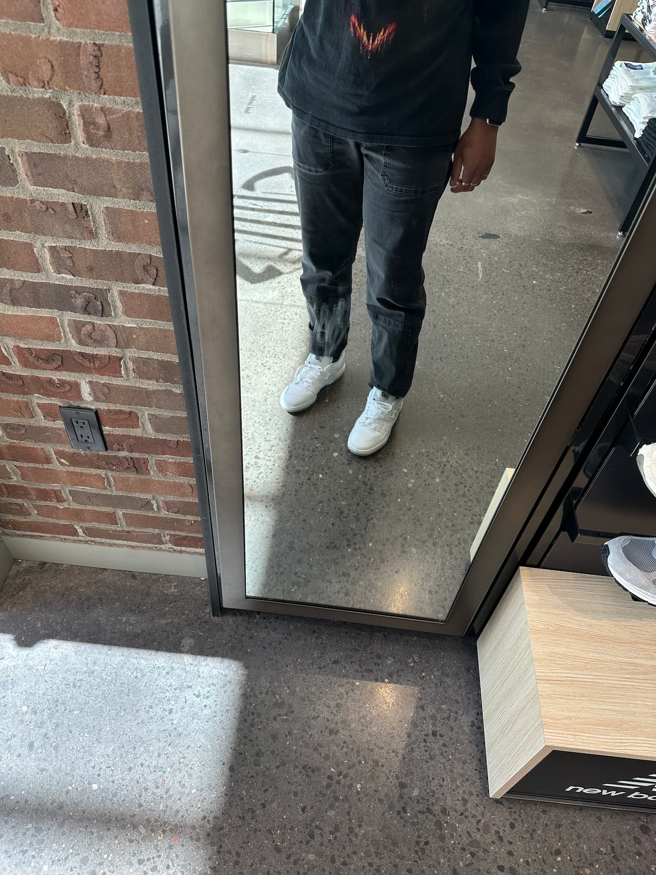 Tristan in dark jeans and white sneakers standing in a doorway between an interior space with polished concrete floors and an exterior brick wall area.