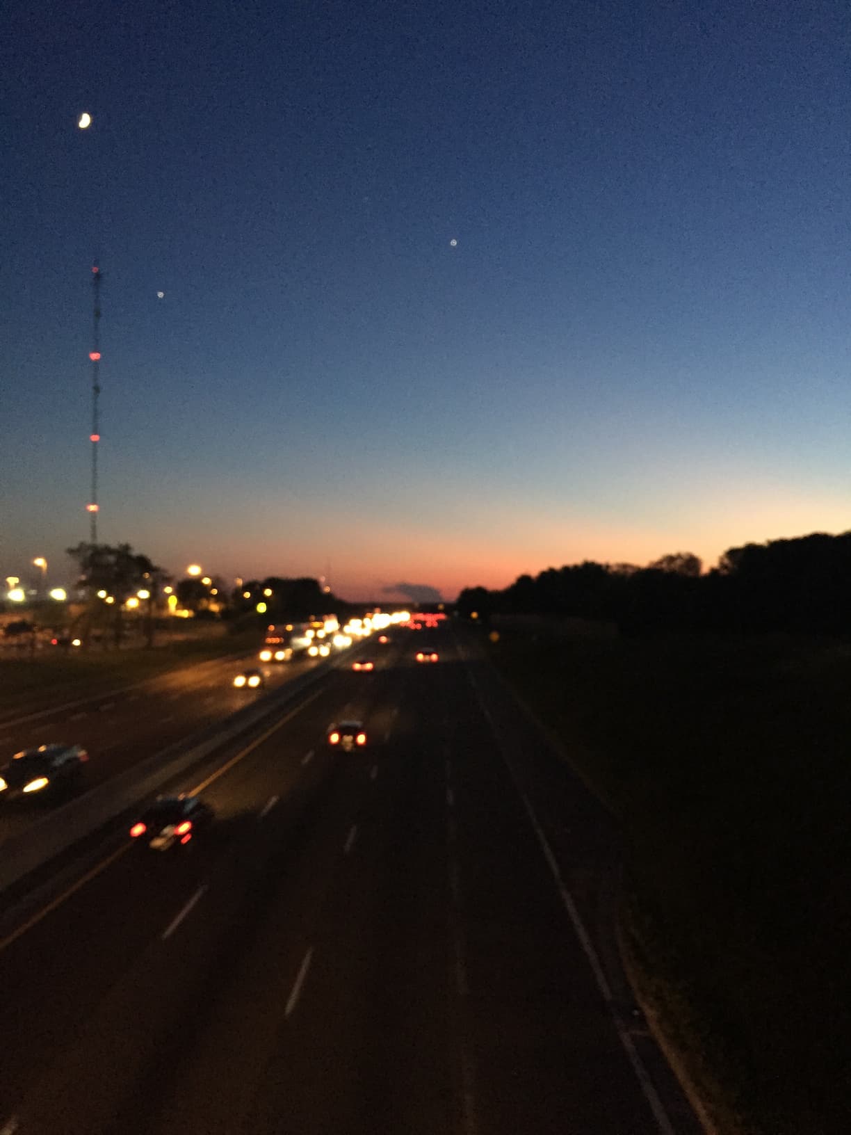 A twilight view of a multi-lane highway with vehicle headlights and taillights creating light trails, set against a gradient sky transitioning from orange near the horizon to deep blue above, with a crescent moon visible and a tall communication tower with red lights on the left.