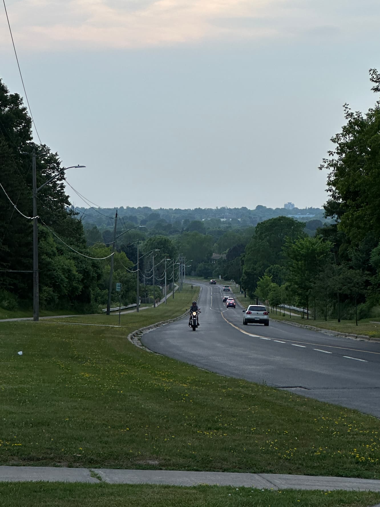 Tree-lined suburban road sloping downhill at dusk, a motorcyclist riding toward the camera with a few cars ahead and power lines along the side under a hazy sky.