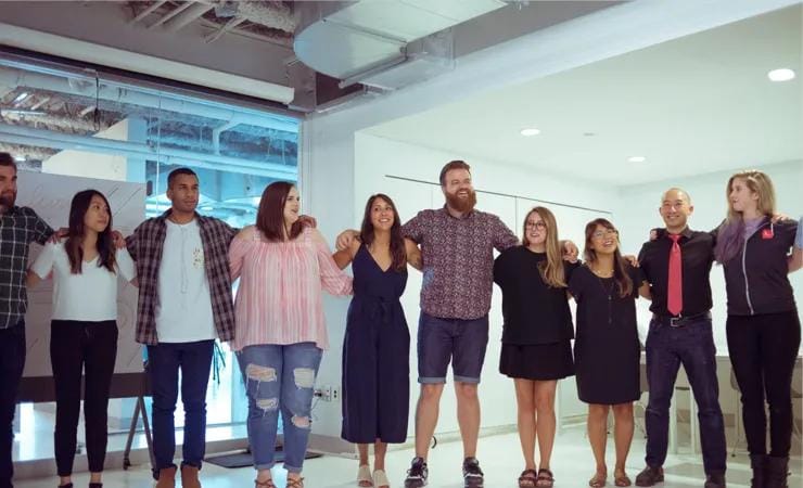 A group of nine people standing in a row inside a modern, brightly lit office space, with their arms around each other’s shoulders, smiling and looking cheerful.