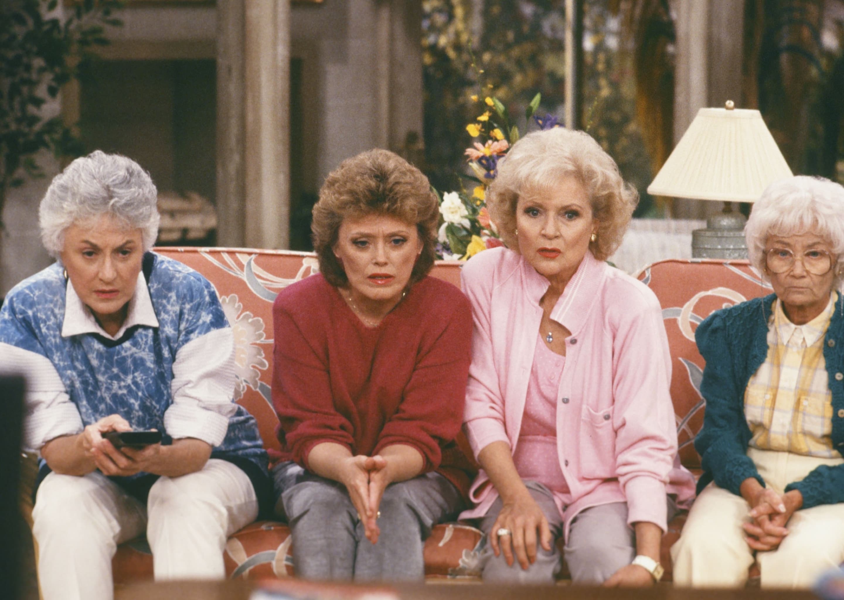 Scene from The Golden Girls TV show featuring the four main characters sitting on a floral couch in their Miami home, wearing colourful 1980s-style clothing.