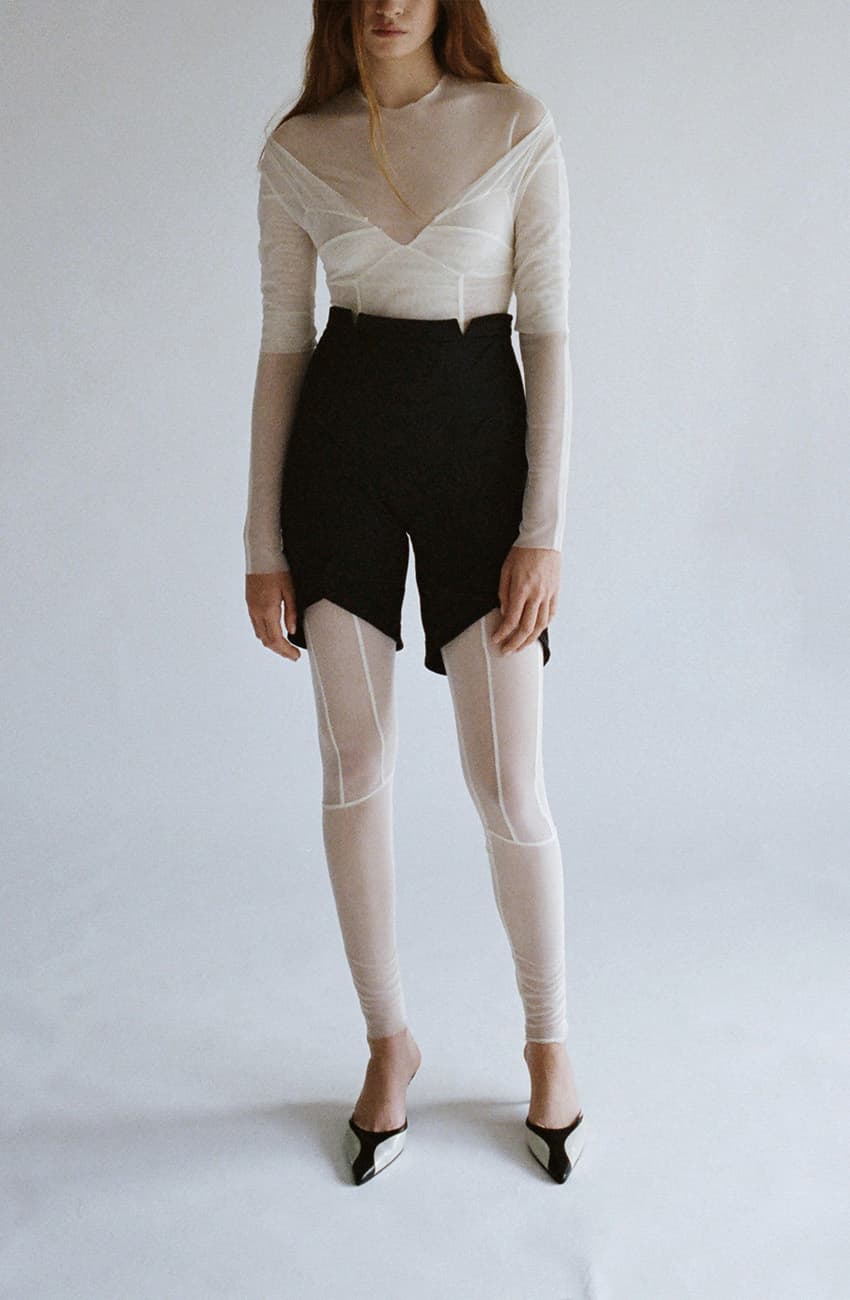 Fashion model wearing minimalist outfit with sheer white top, black structured shorts, white leggings and black pointed heels against neutral background.