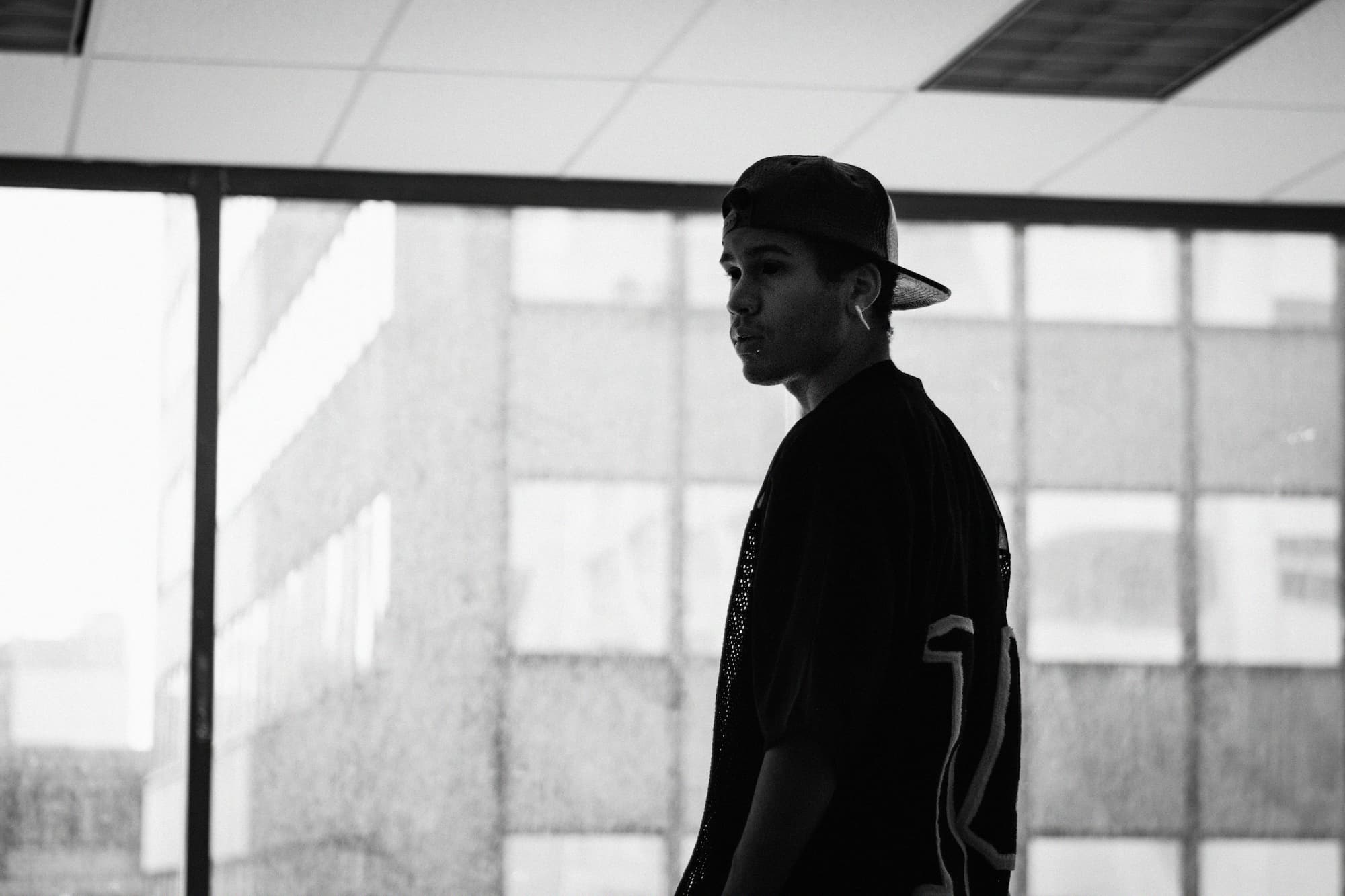 Black and white profile silhouette of a young person wearing a backwards cap in an indoor space with large windows and drop ceiling, creating a contemplative urban atmosphere.