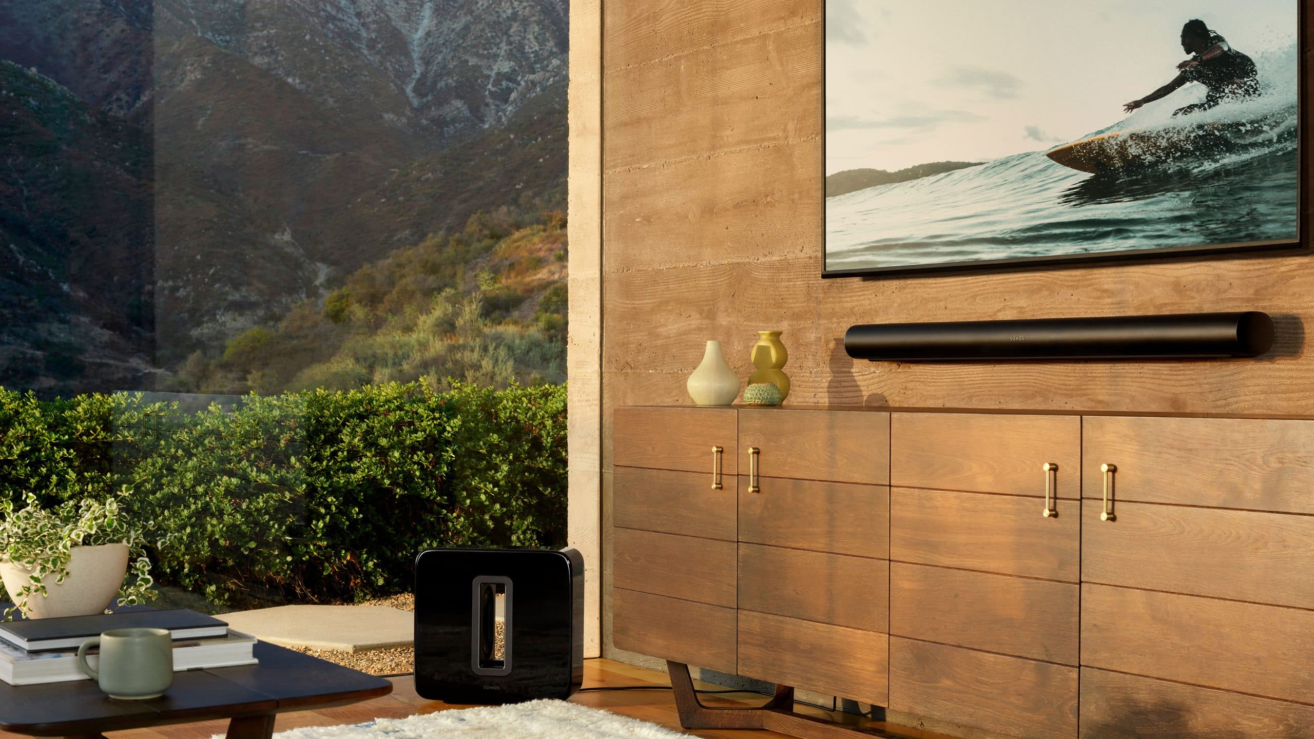 Modern living room with wooden media console, soundbar, and surfing artwork, featuring mountain views through large windows and Sonos speaker on coffee table.